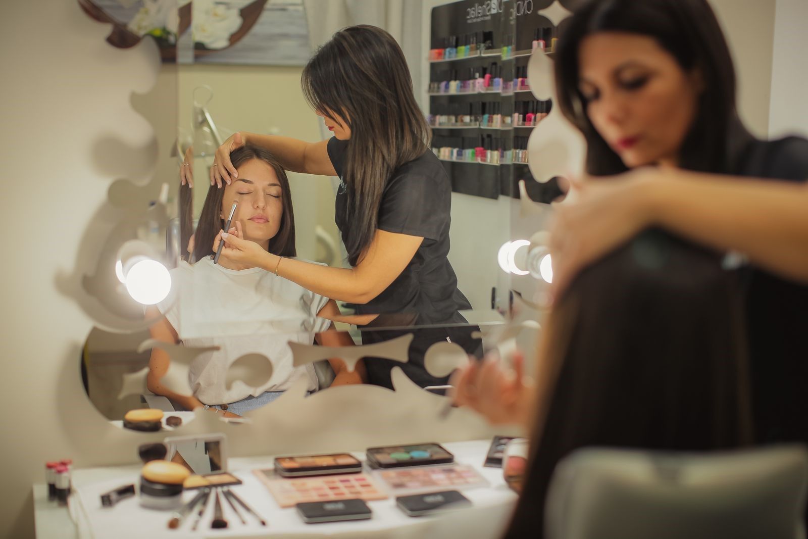 Una truccatrice applica il trucco a una donna davanti a uno specchio da trucco, con i cosmetici sparsi sul tavolo.