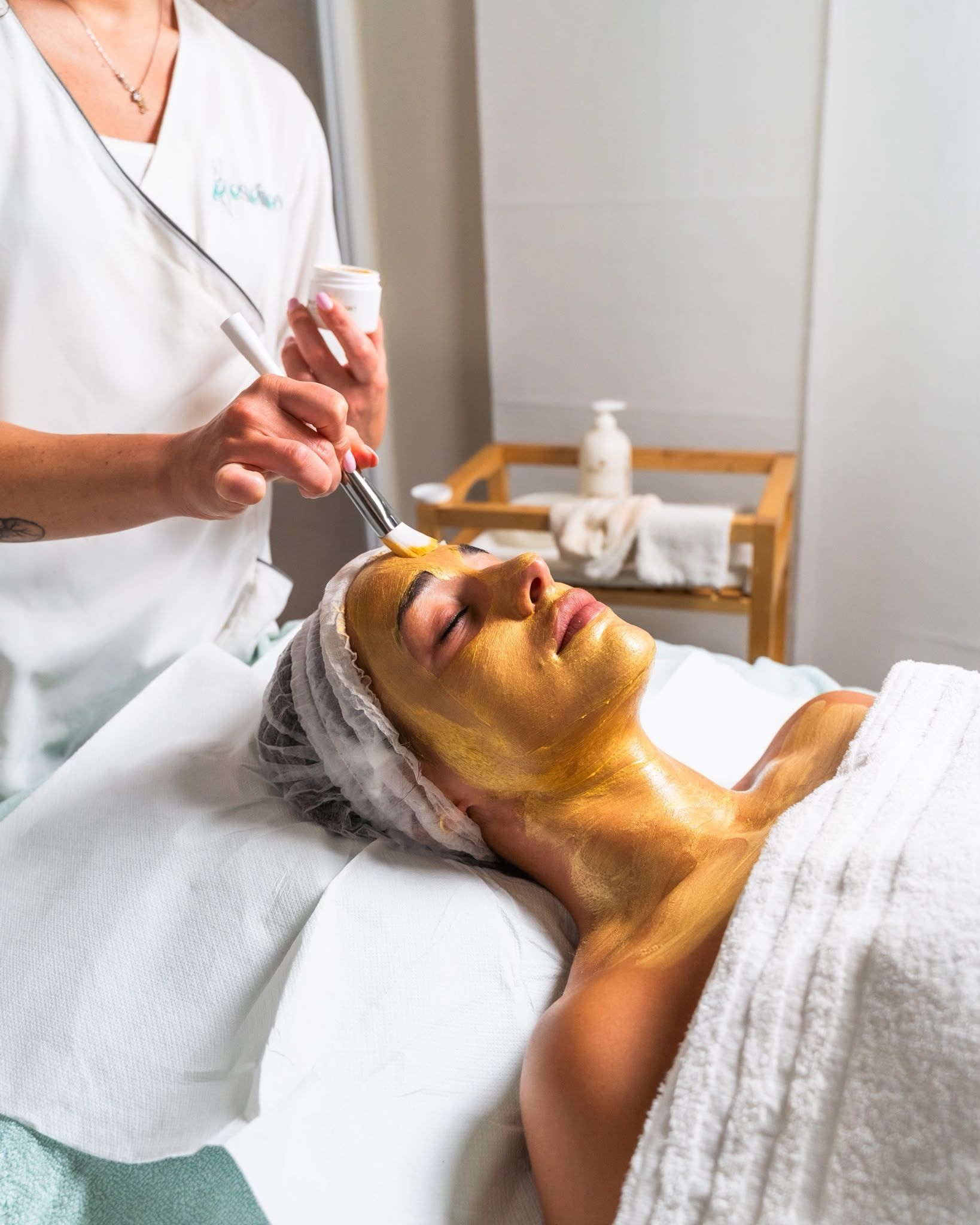 Un'estetista applica un trattamento a una cliente sdraiata su un lettino da spa con una maschera viso dorata.