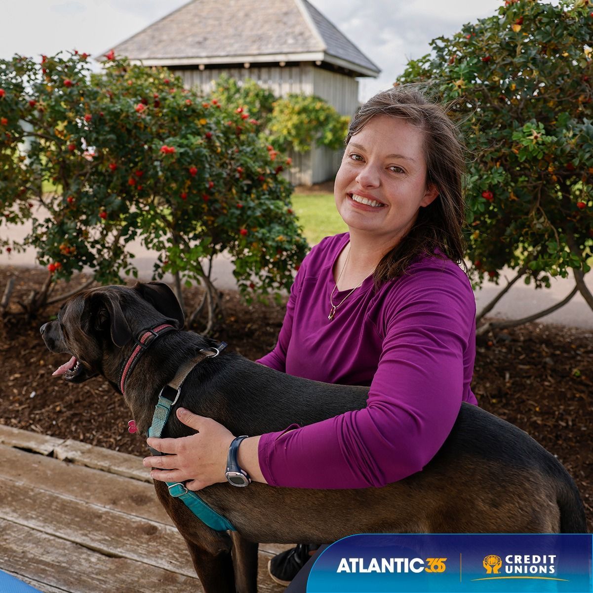 Smiling woman in a purple top hugging a black dog outdoors near bushes and a building