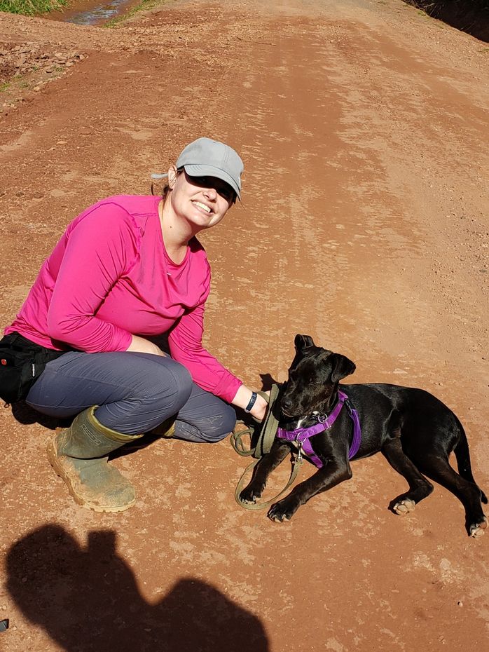Person kneeling with a black dog on a leash on a dirt path, both facing the camera.