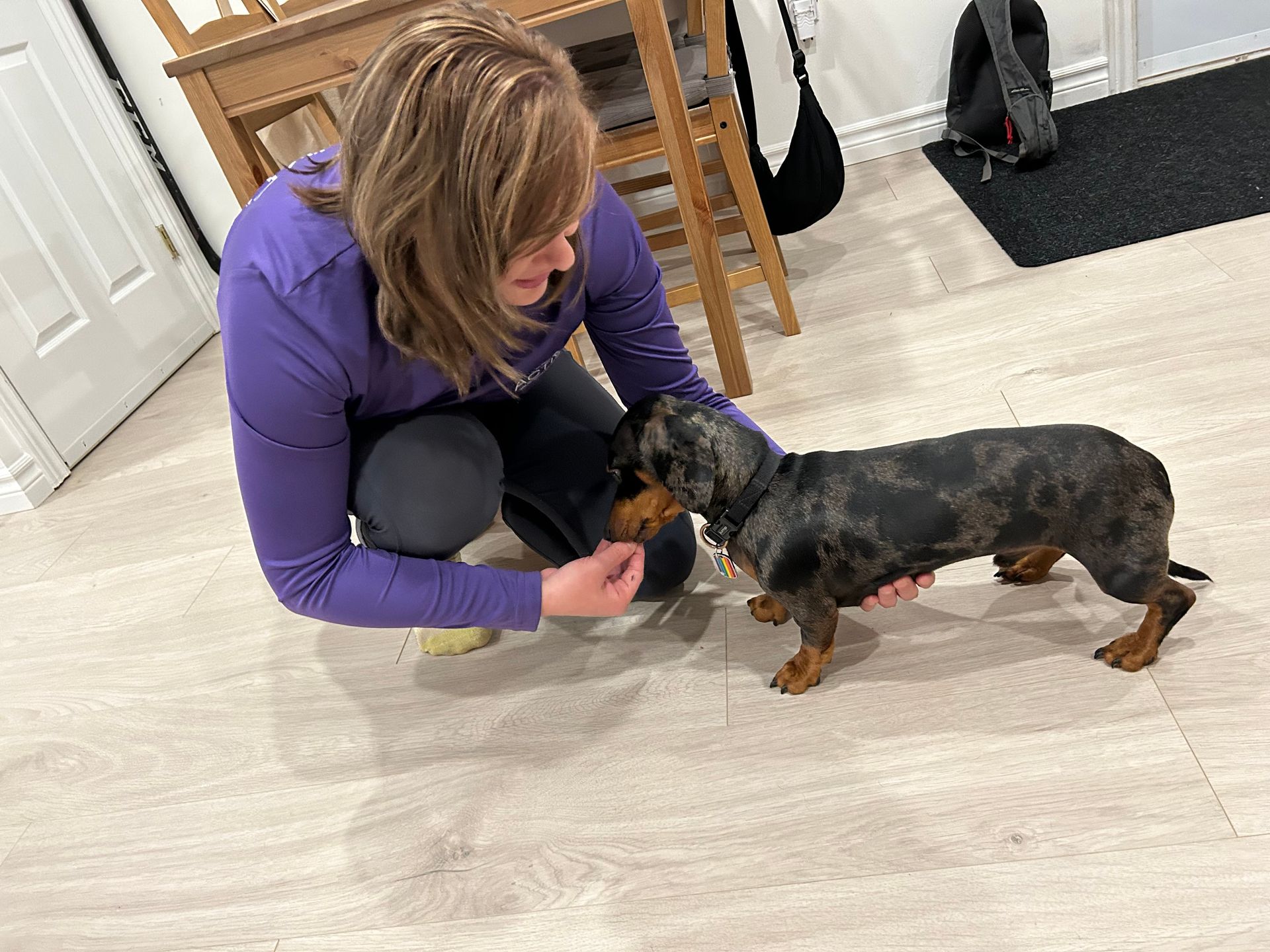 Woman crouches indoors, offering treats to a small black-and-brown dog on a light floor.