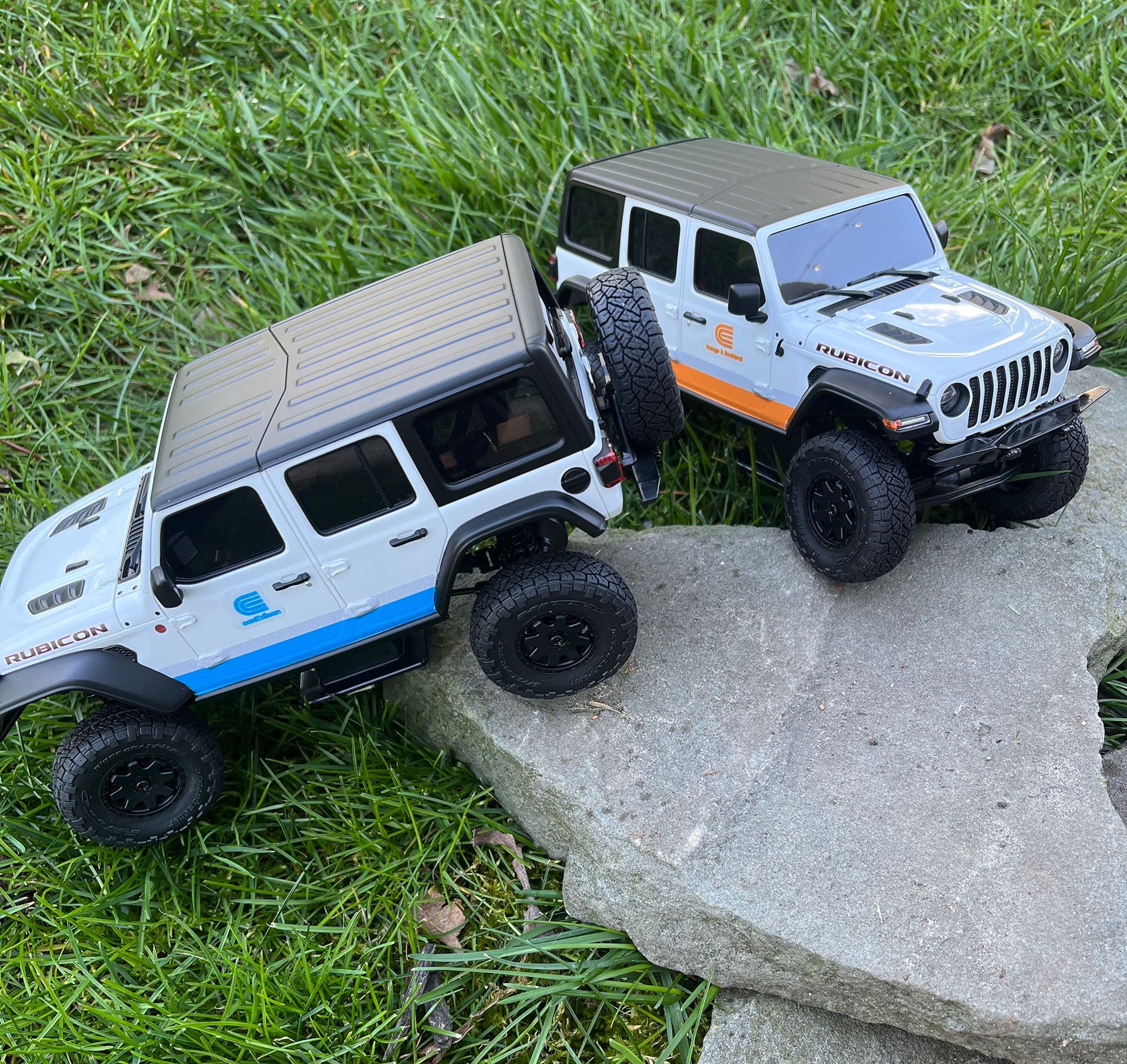 Two toy off-road trucks on grass and a rock, one white with blue accents and one gray with orange accents.