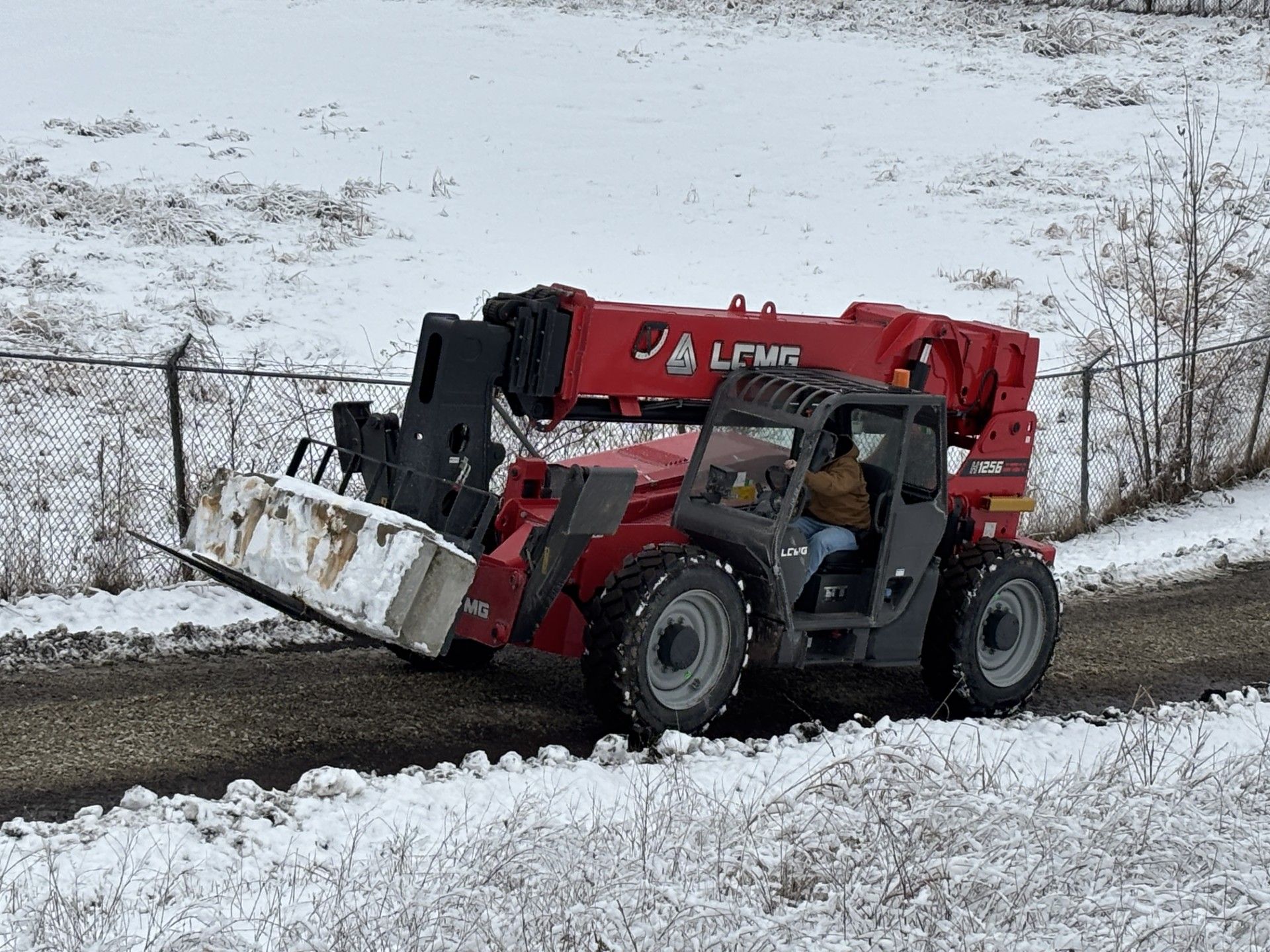 Red telehandler with bucket on snowy road, person driving, near a fence.