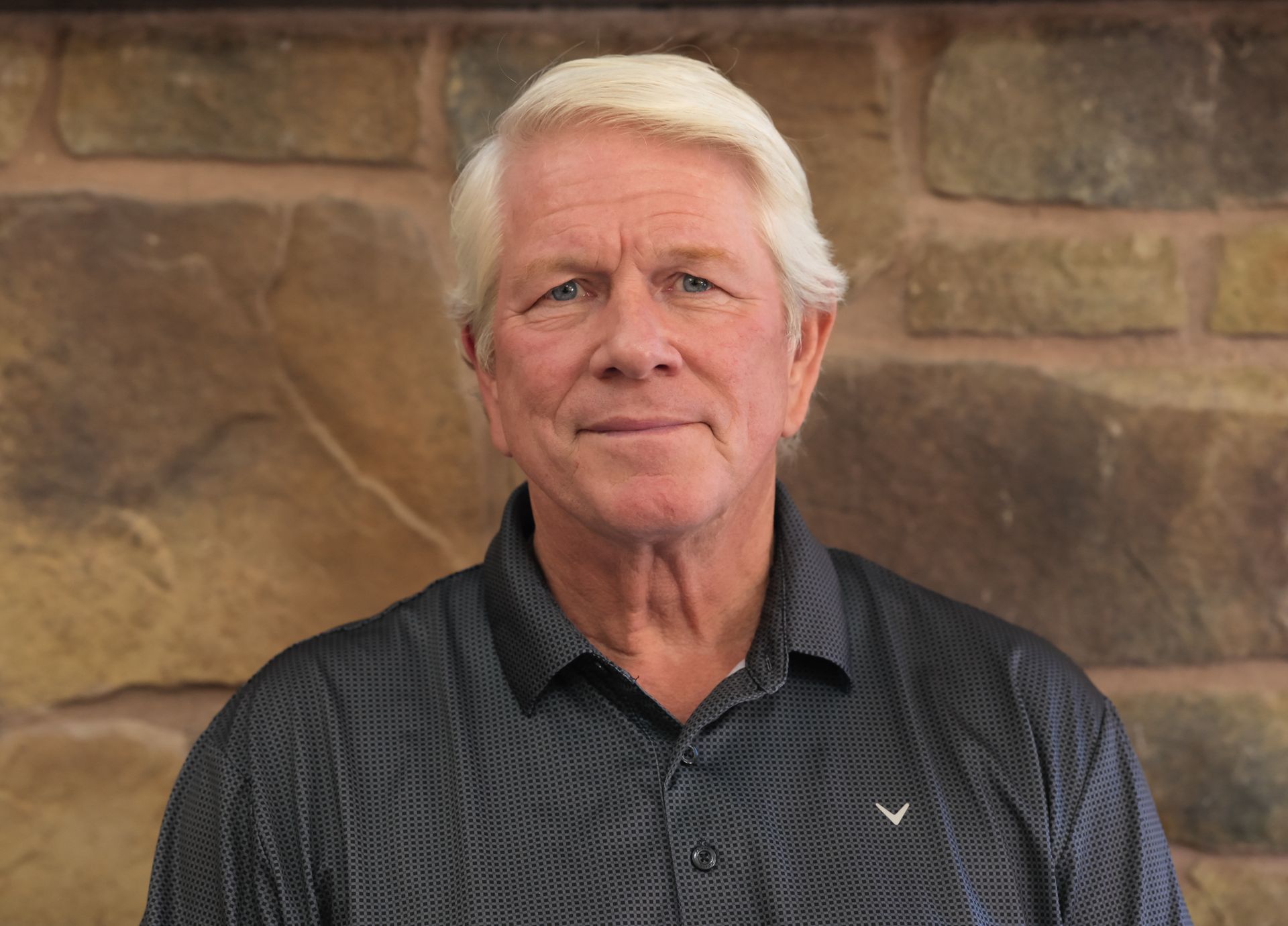 Man with white hair, wearing a dark patterned shirt, stands in front of a brick wall.