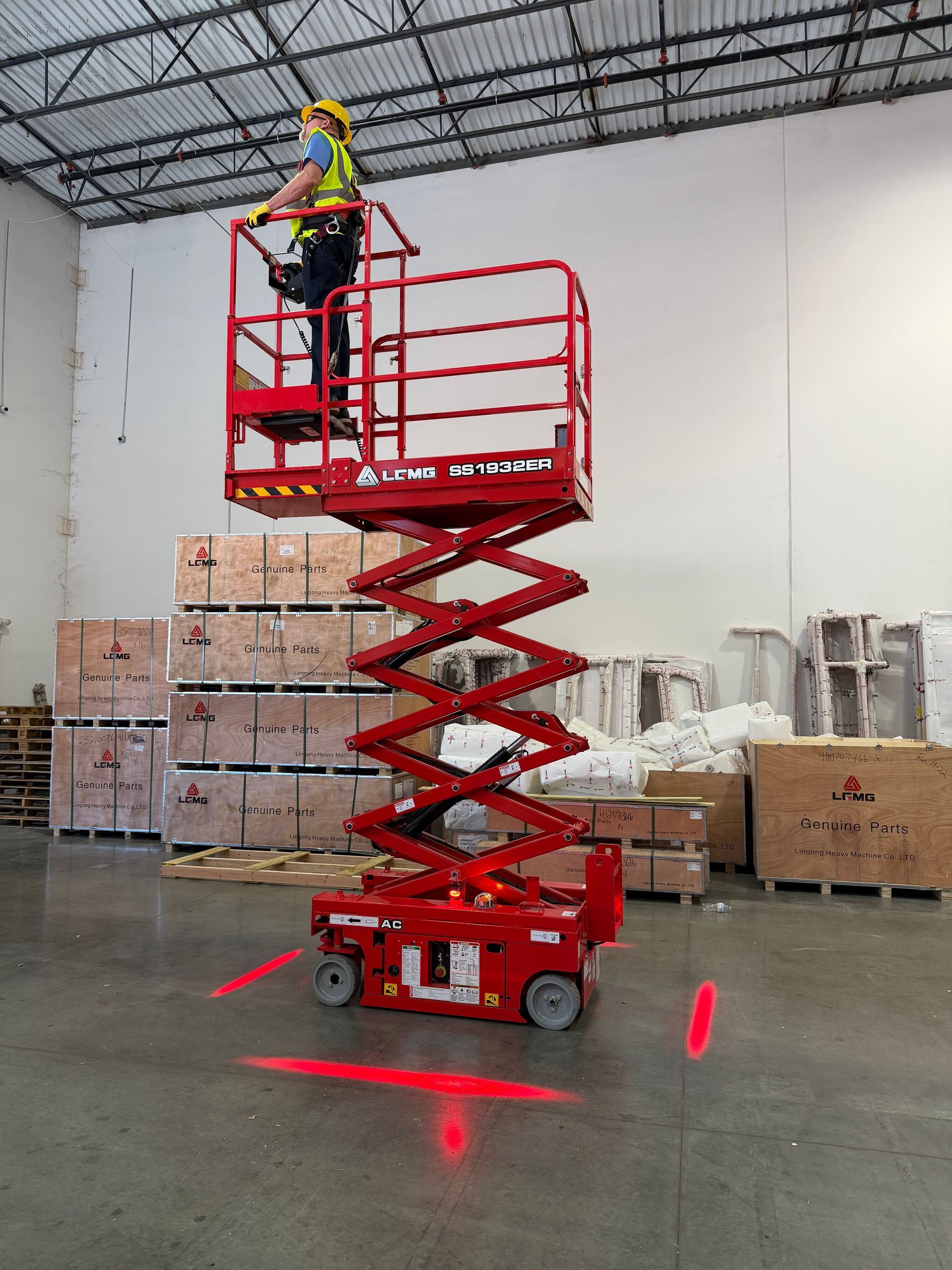 Worker on red scissor lift in warehouse, wearing a hard hat and safety vest.