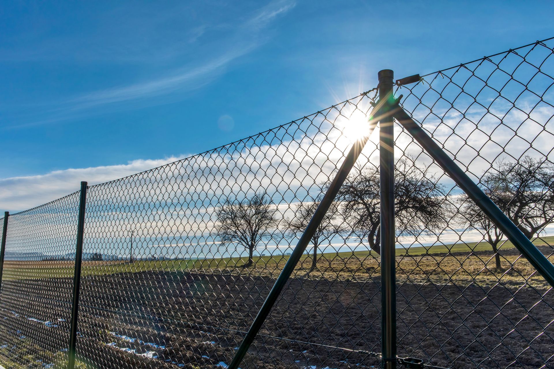 The sun is shining through a chain link fence in a field.