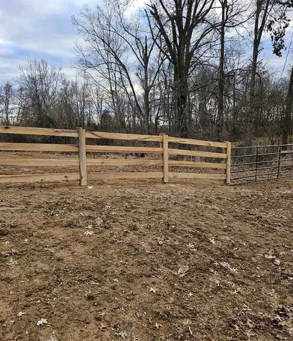 A wooden fence surrounds a dirt field with trees in the background.
