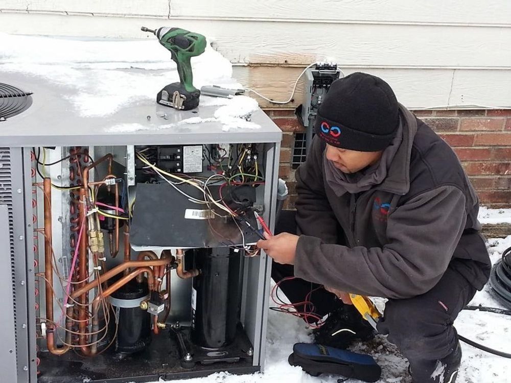 Technician repairing an open HVAC unit, using a drill beside exposed wiring and copper pipes outdoors
