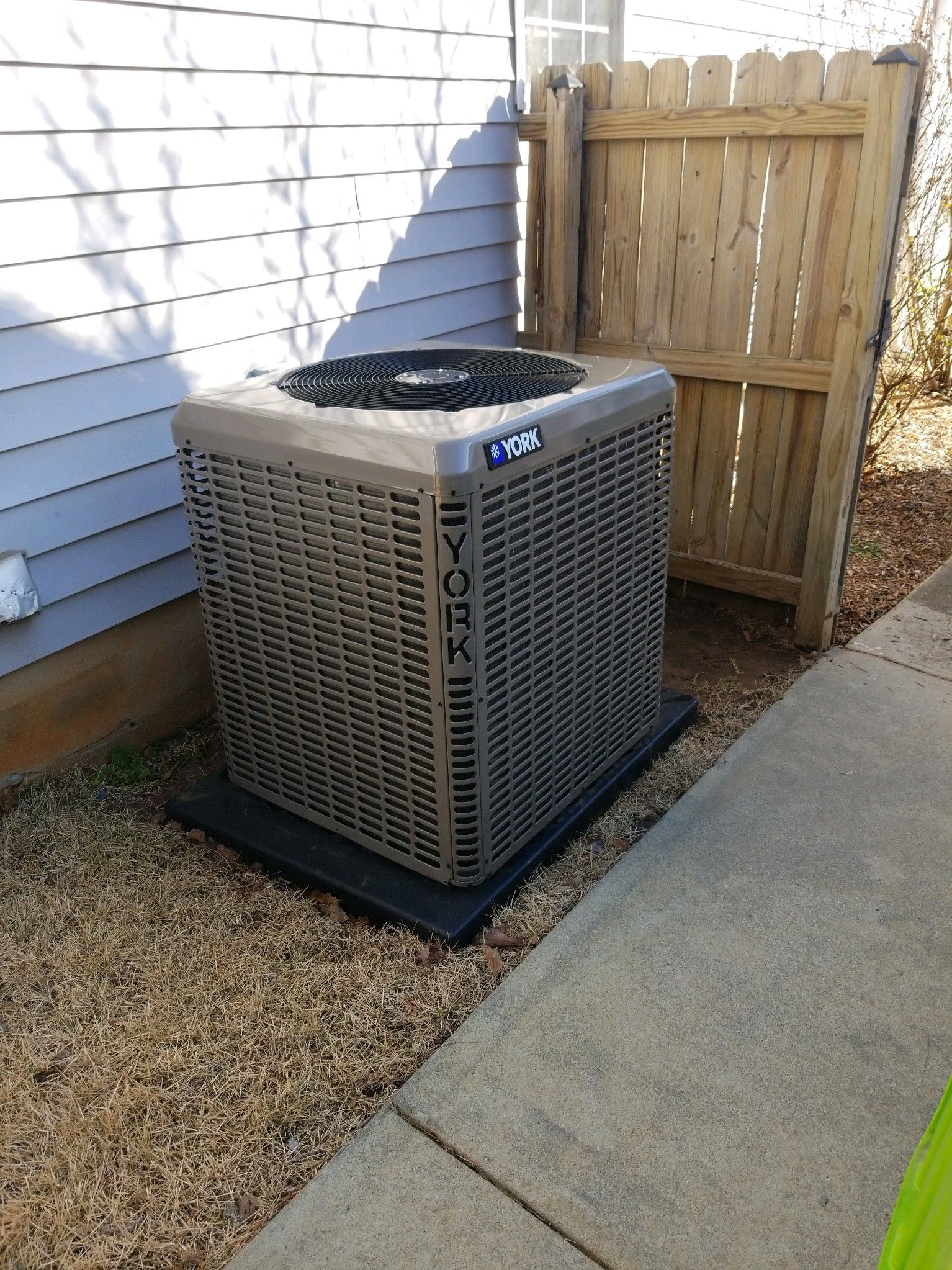 Outdoor air conditioner unit beside a house and wooden fence on a gravel-and-concrete patio.