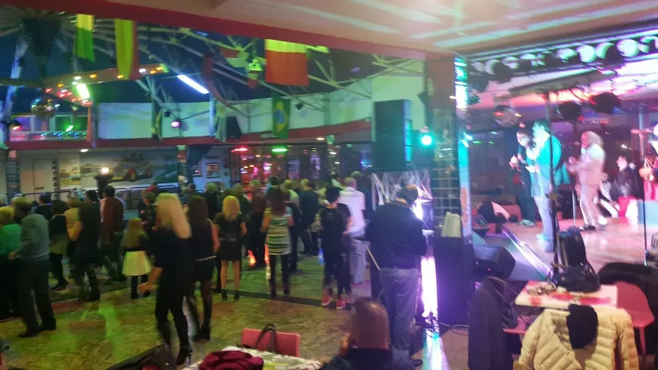 Folla in un vivace locale al chiuso, con persone che ballano e un artista sul palco sotto luci brillanti e bandiere colorate.