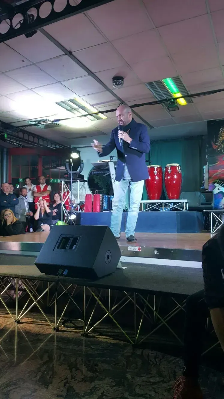 Una persona con un blazer blu e jeans chiari parla al microfono su un palco di fronte a due conga rosse.