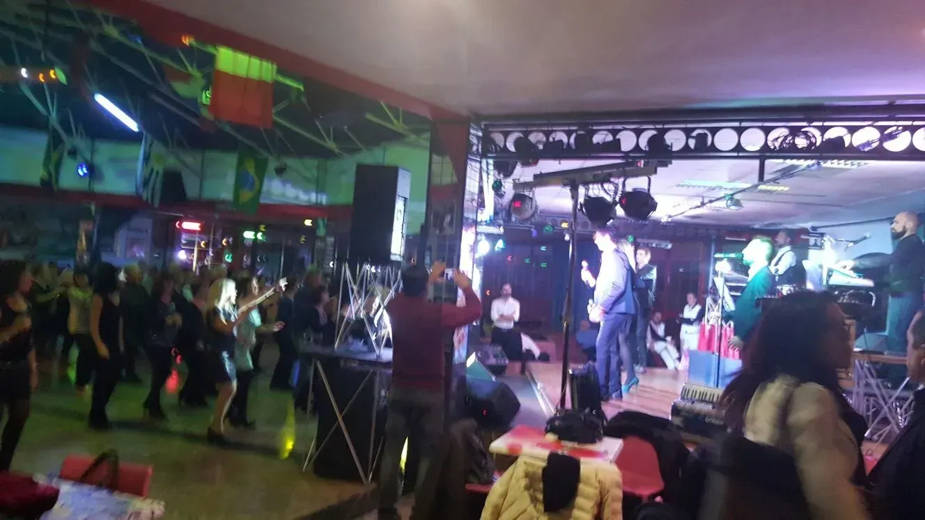 In un ambiente festoso e scarsamente illuminato da luci colorate, alcune persone ballano su una pista da ballo di fronte a un palco con degli artisti.