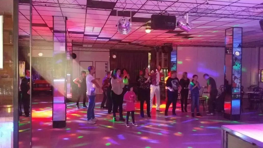 Un gruppo di persone in piedi in una sala da ballo colorata e scarsamente illuminata, decorata con colonne a specchio e luci da discoteca.