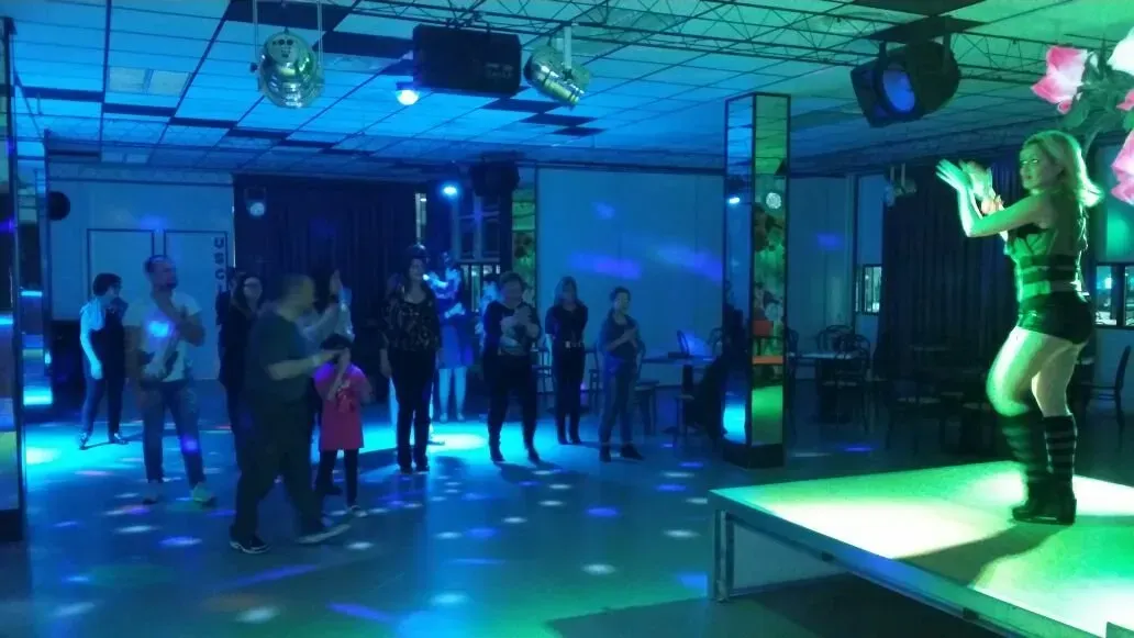 Una donna balla su un palco in un locale scarsamente illuminato da luci blu e verdi, mentre diverse persone la osservano dalla pista da ballo.