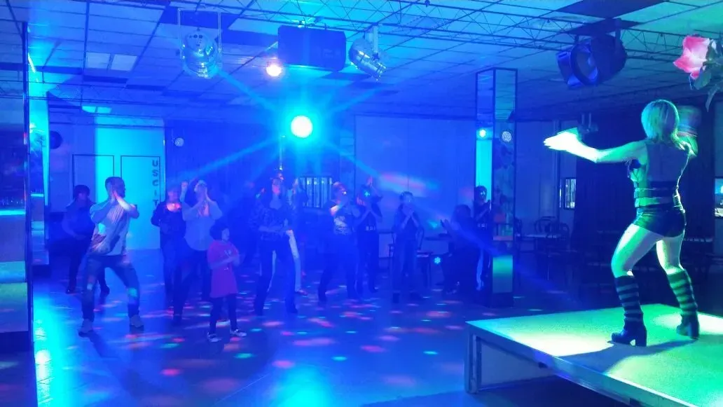 Una persona balla su un palco in un locale illuminato di blu, mentre la folla la guarda e balla sulla pista da ballo.