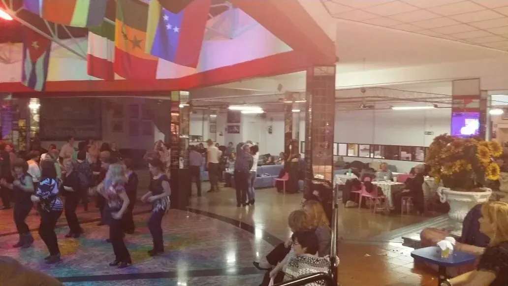 In una sala addobbata con bandiere, alcune persone ballano, mentre altre siedono ai tavoli in uno spazio interno illuminato a giorno.