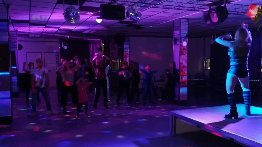 Una persona vestita di scuro si esibisce su un palco in un locale notturno scarsamente illuminato e dalle tonalità violacee, mentre un piccolo gruppo balla nelle vicinanze.