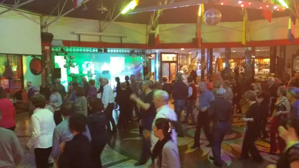 Una folla eterogenea di persone balla su un pavimento di legno in un locale scarsamente illuminato da luci di scena verdi e una palla da discoteca.