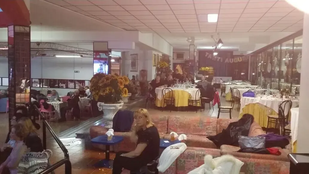 Una sala da pranzo interna scarsamente illuminata con tavoli e sedie sparsi e persone che si rilassano su comode poltrone.