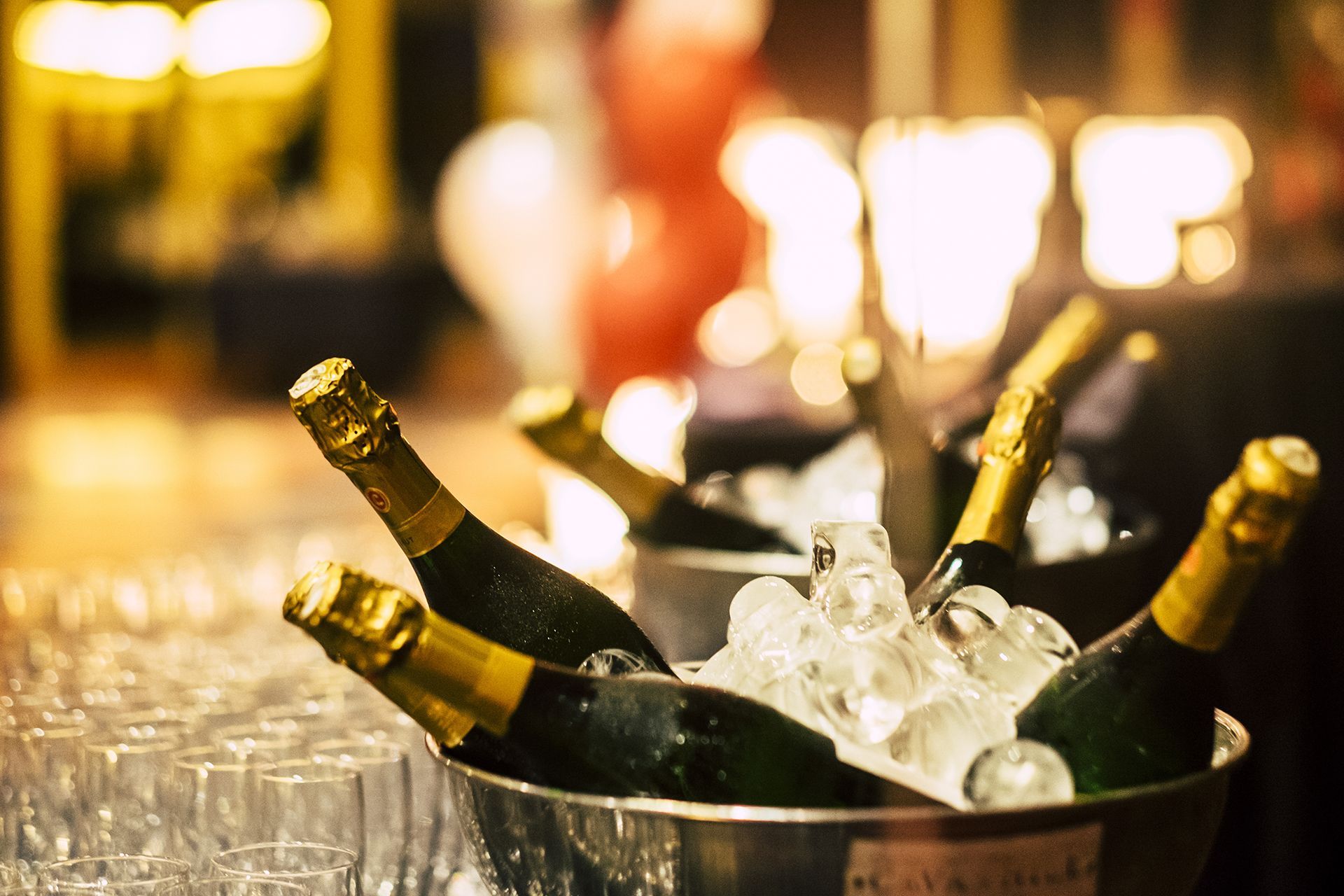 Diverse bottiglie di champagne in un secchiello del ghiaccio, con molti bicchieri vuoti nelle vicinanze, in un'atmosfera festosa e a luci soffuse.