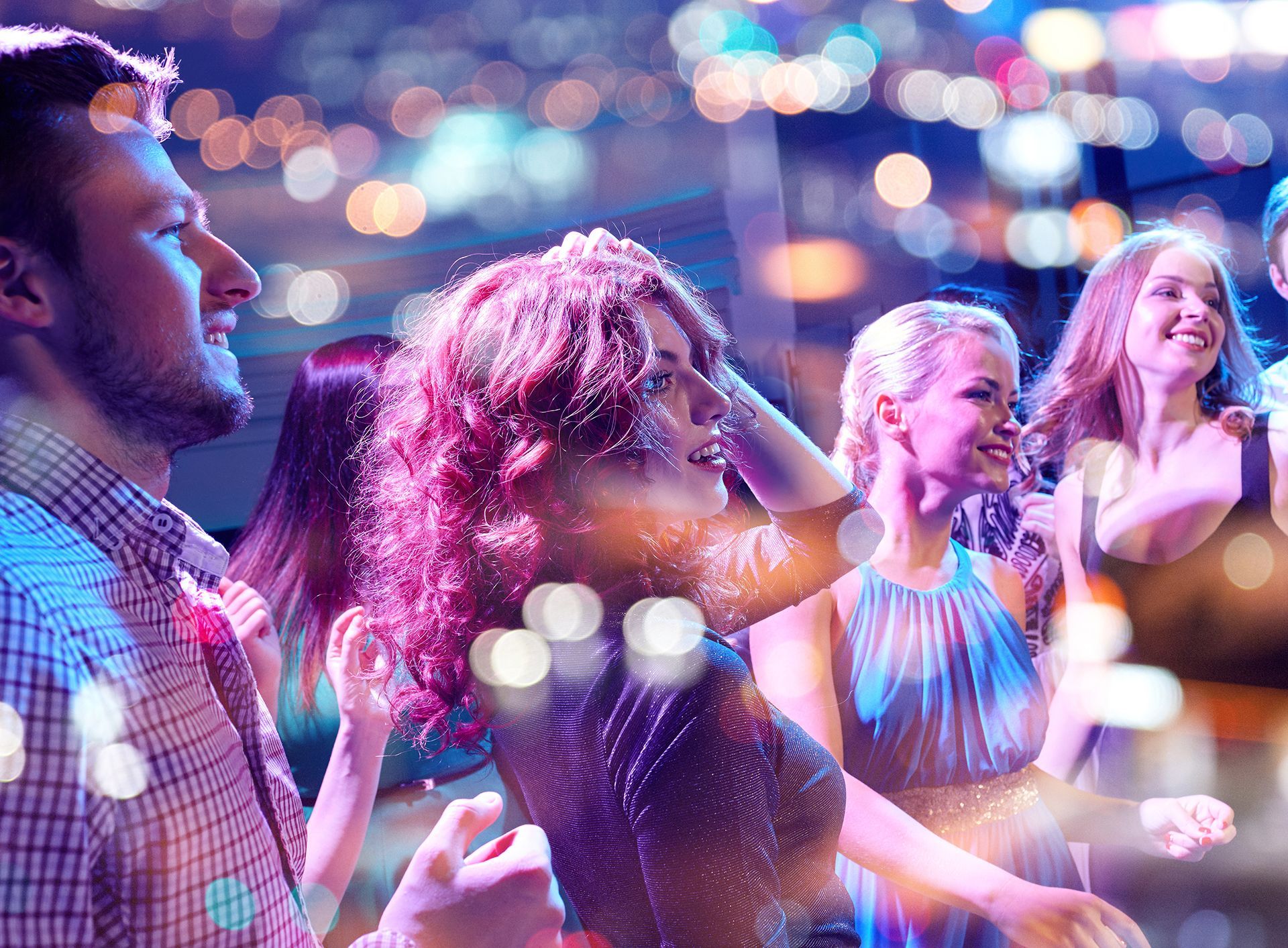 In un locale notturno illuminato da luci soffuse e colorate, le persone ballano e sorridono.