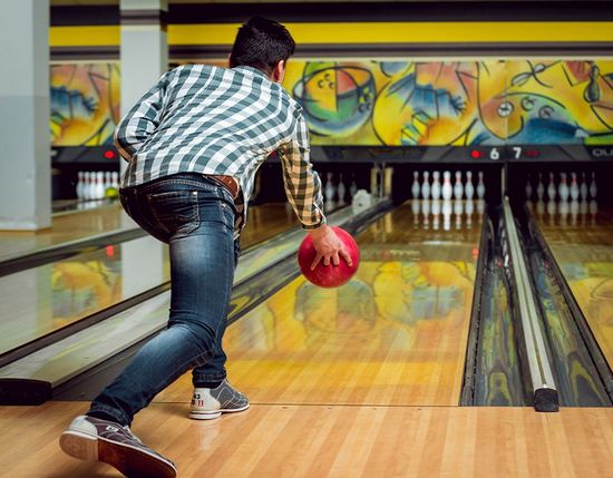 In una sala da bowling, una persona con una camicia a quadri lancia una palla da bowling rossa lungo una pista, indirizzandola verso i birilli.
