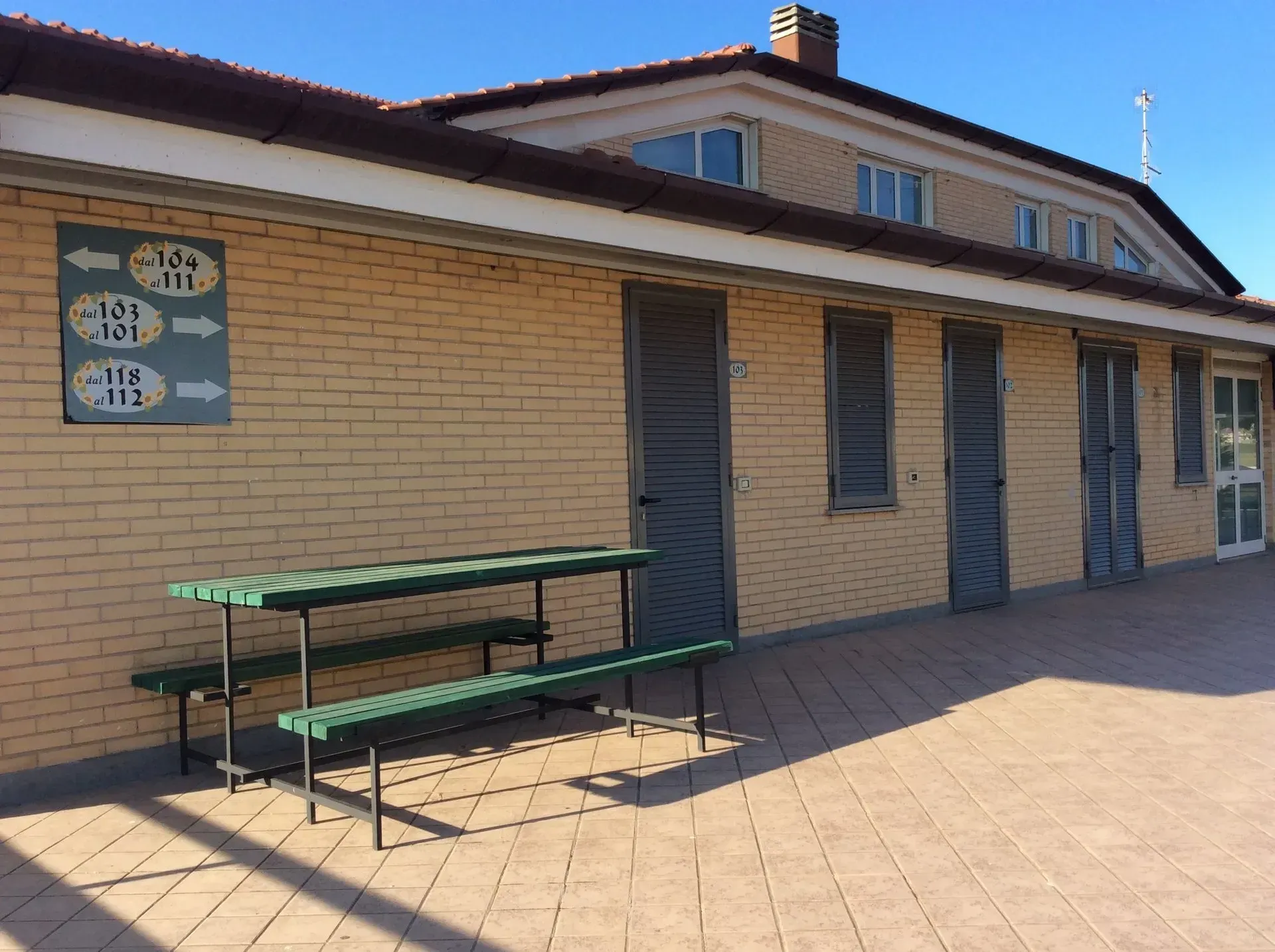 Un edificio in mattoni con diverse porte e un tavolo da picnic verde su un patio pavimentato, con segnaletica direzionale sulla parete.