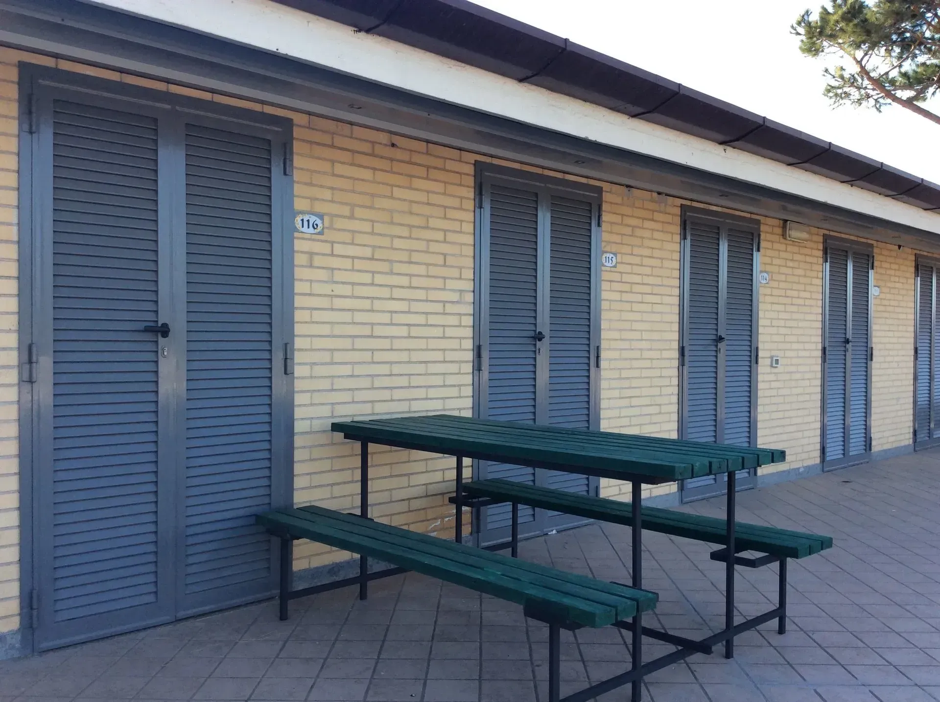 Un tavolo da picnic verde si trova di fronte a un edificio in mattoni gialli con una serie di porte a persiana grigie su un patio piastrellato.