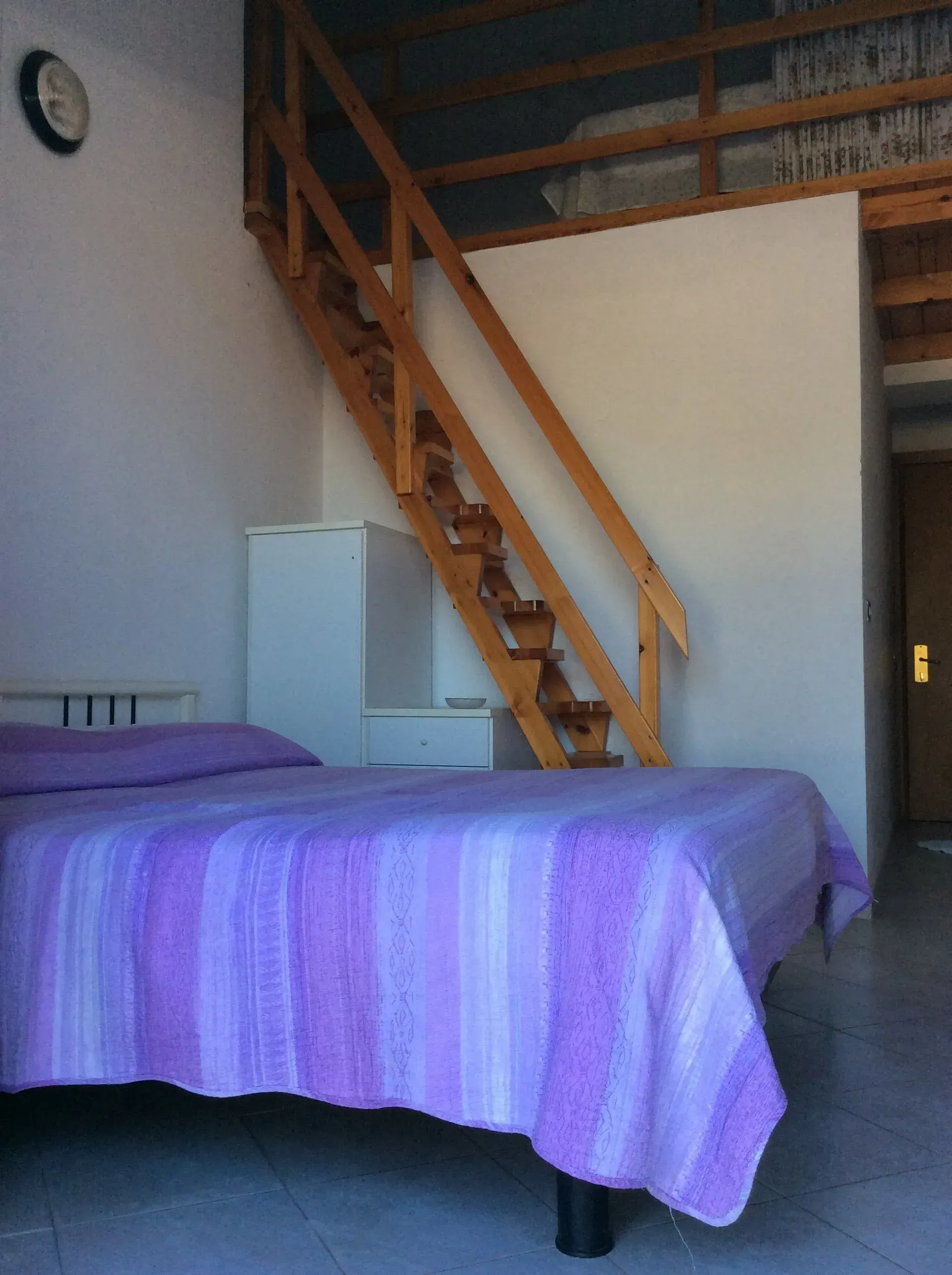 Una camera da letto con un letto con una trapunta a righe viola, una cassettiera bianca e una scala in legno che conduce a un soppalco.