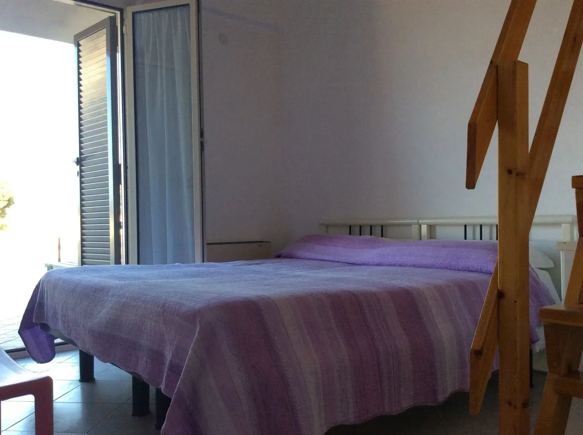Una camera da letto con un letto matrimoniale coperto da una trapunta a righe viola, accanto a una scala di legno e a una porta che conduce all'esterno.