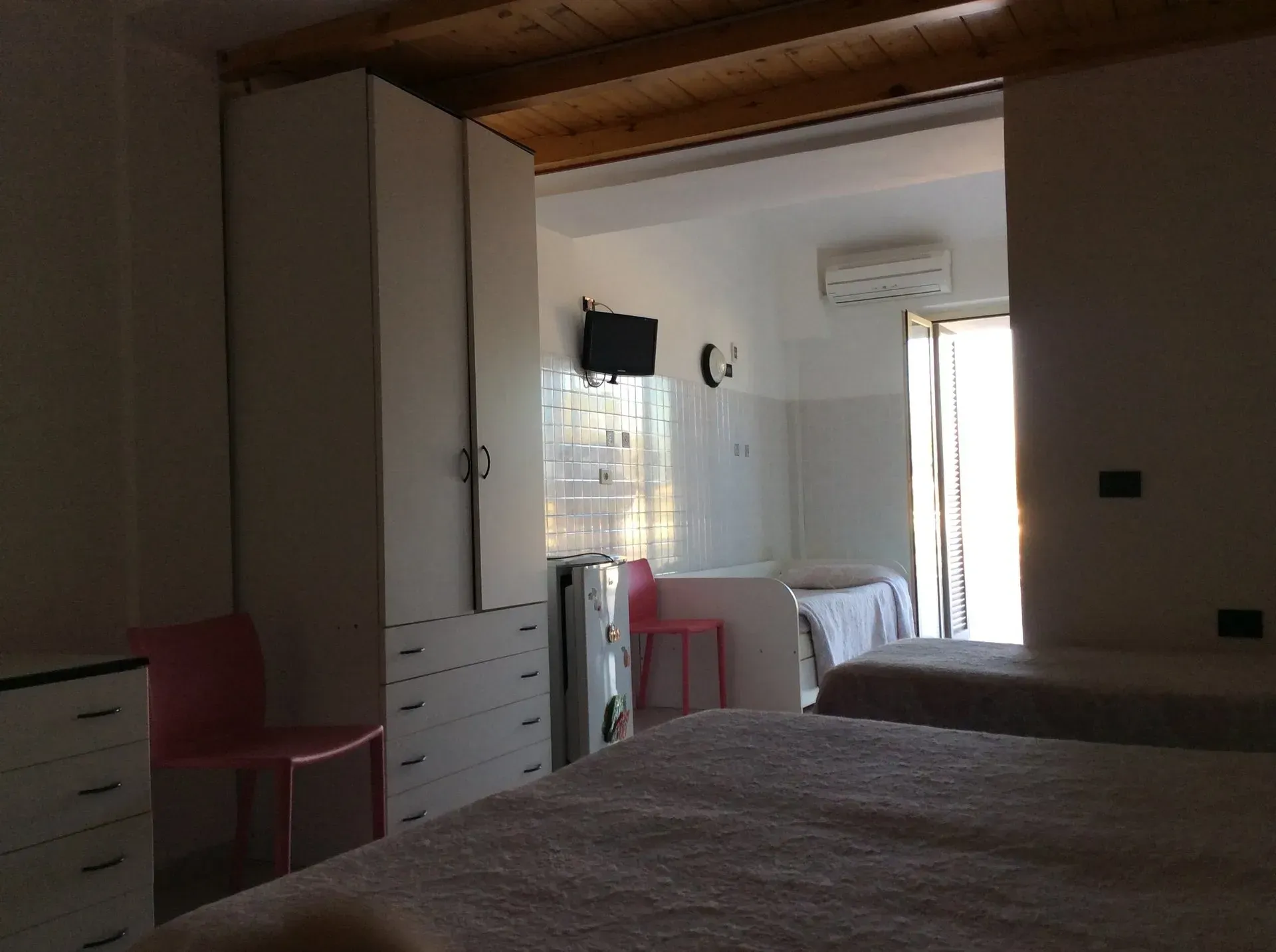 Una camera d'albergo con due letti, un armadio bianco, una sedia rosa, pareti piastrellate e una porta aperta che conduce a una zona luminosa.
