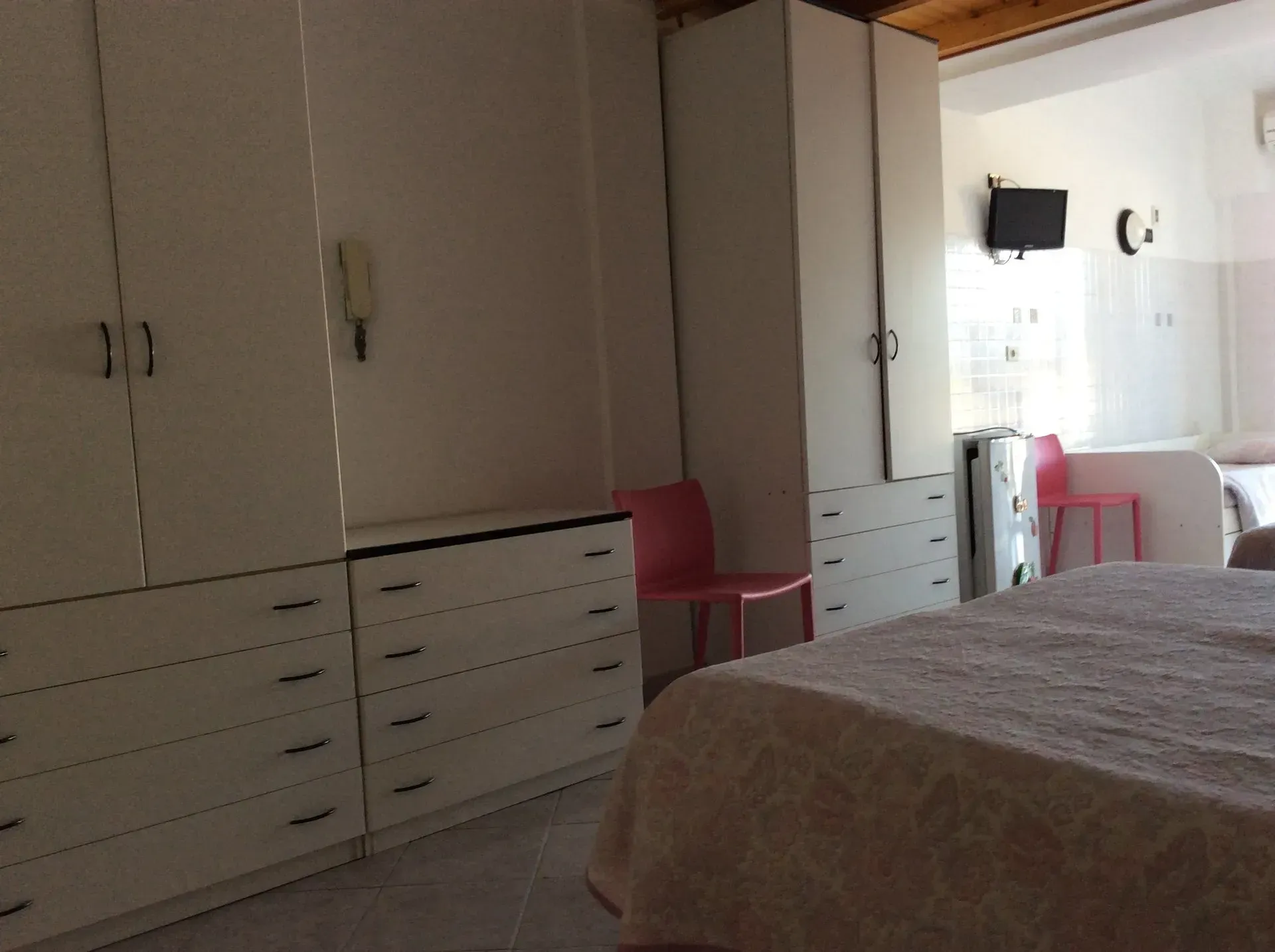 Una camera da letto con armadi e cassettiera bianchi, una sedia rossa, un letto con una coperta a fantasia e un televisore a parete.