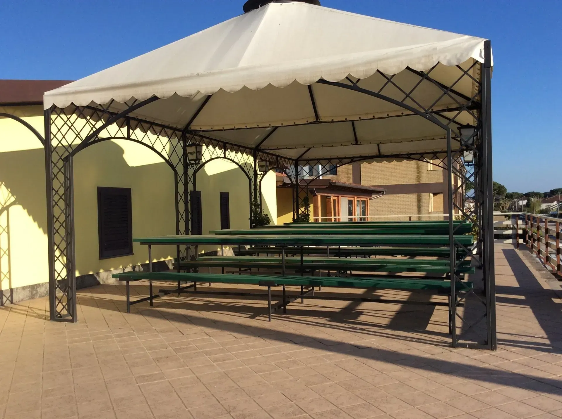 Un gazebo in metallo con una copertura color crema ombreggia lunghi tavoli e panchine in legno verde su un patio pavimentato.