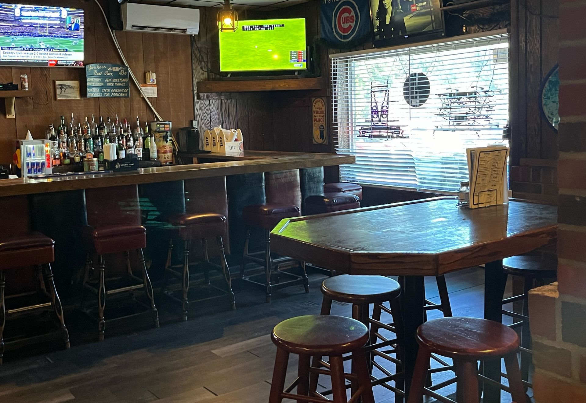 Interior of a bar with a wooden counter and stools, tables, and TV screens playing sports.
