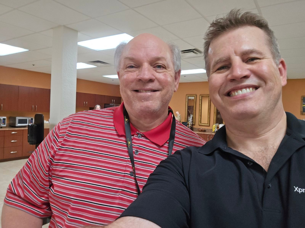 Two men smiling and posing for a selfie in an office breakroom with orange walls and wooden cabinets.