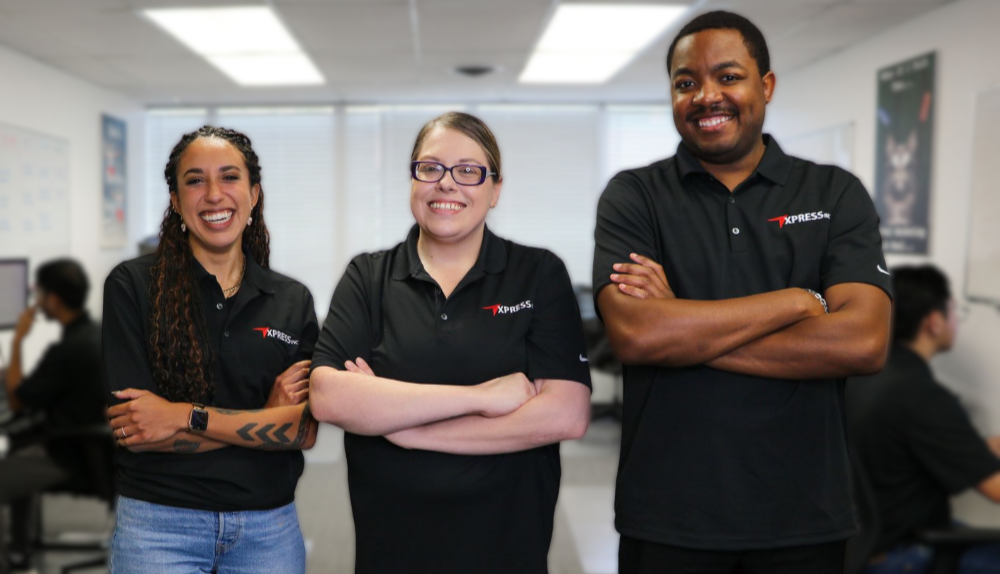 Three colleagues in branded black polo shirts stand smiling with arms crossed in an office setting.