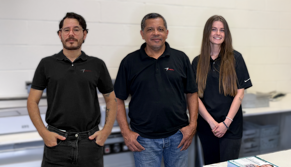 Three people in black polo shirts stand side by side in a workspace with a white wall and office equipment behind them.