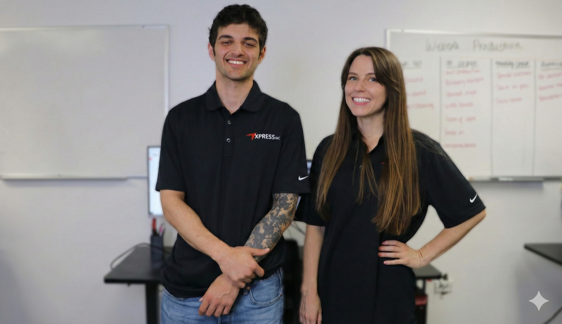 Two people in black polo shirts stand smiling side-by-side in an office with whiteboards in the background.