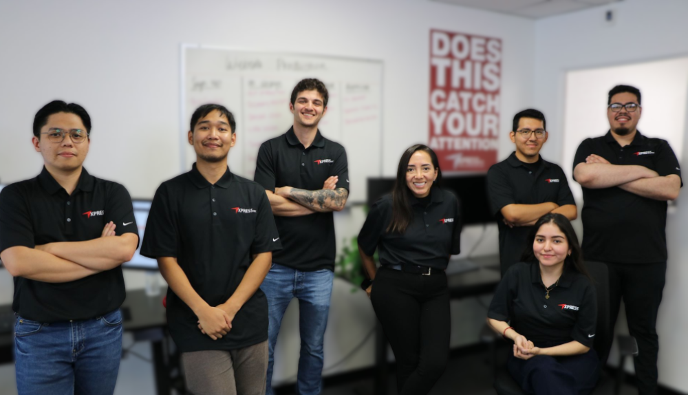 A team of seven professionals in branded black polo shirts stands in an office in front of a whiteboard and wall poster.