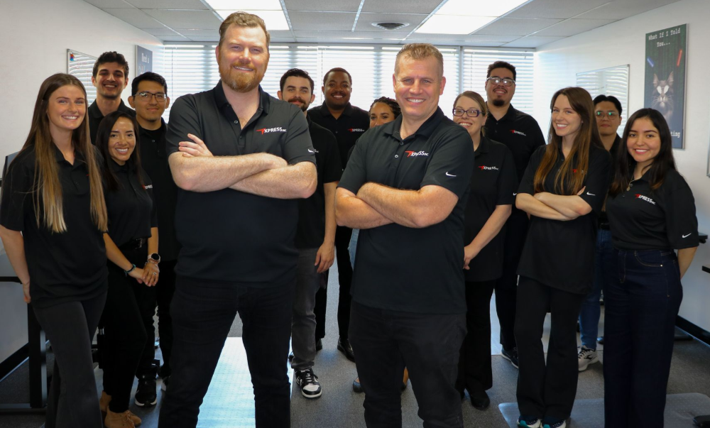 Two men in black shirts with arms crossed, standing in an office with other people working at computers.