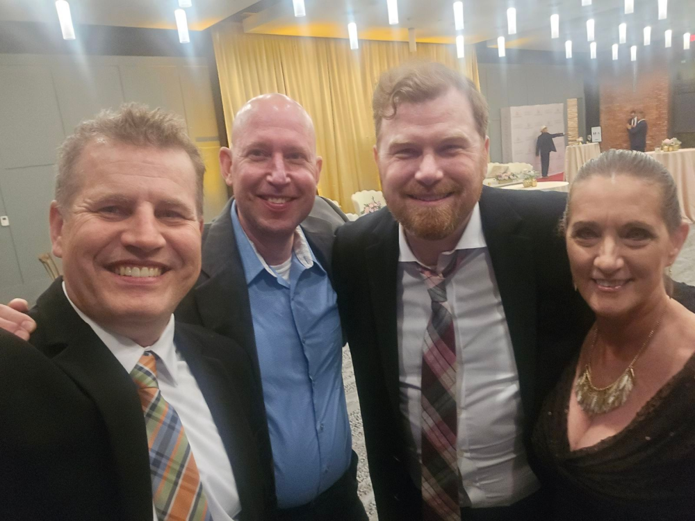 Four people smiling in a well-lit room, possibly a formal event. Two men wear suits, one a patterned tie. A woman has a necklace, wearing a dark top.