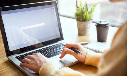 A person using a laptop on a wooden desk, with a small potted plant and a coffee cup nearby.