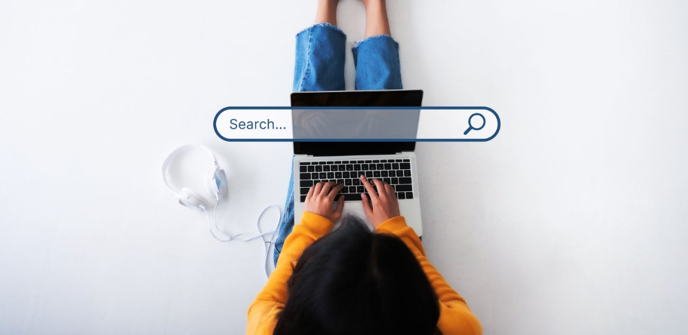 A person in a yellow sweater sits on the floor using a laptop with a digital search bar overlay.