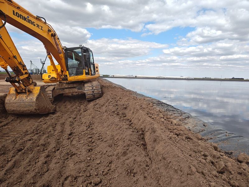 Una excavadora amarilla construyendo un dique de contención de barro junto a una orilla tranquila bajo un cielo nublado