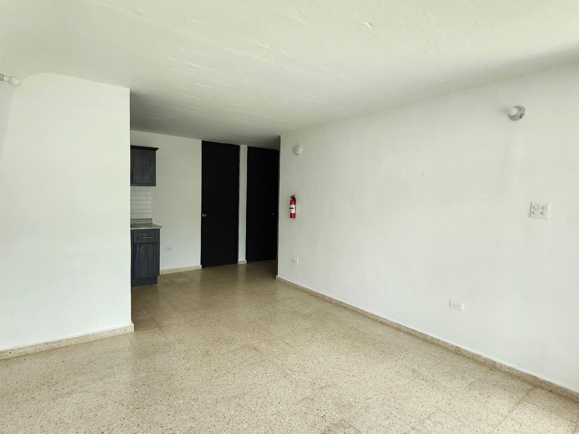 Empty white room with speckled floor, dark doorway, and a small kitchen area in back