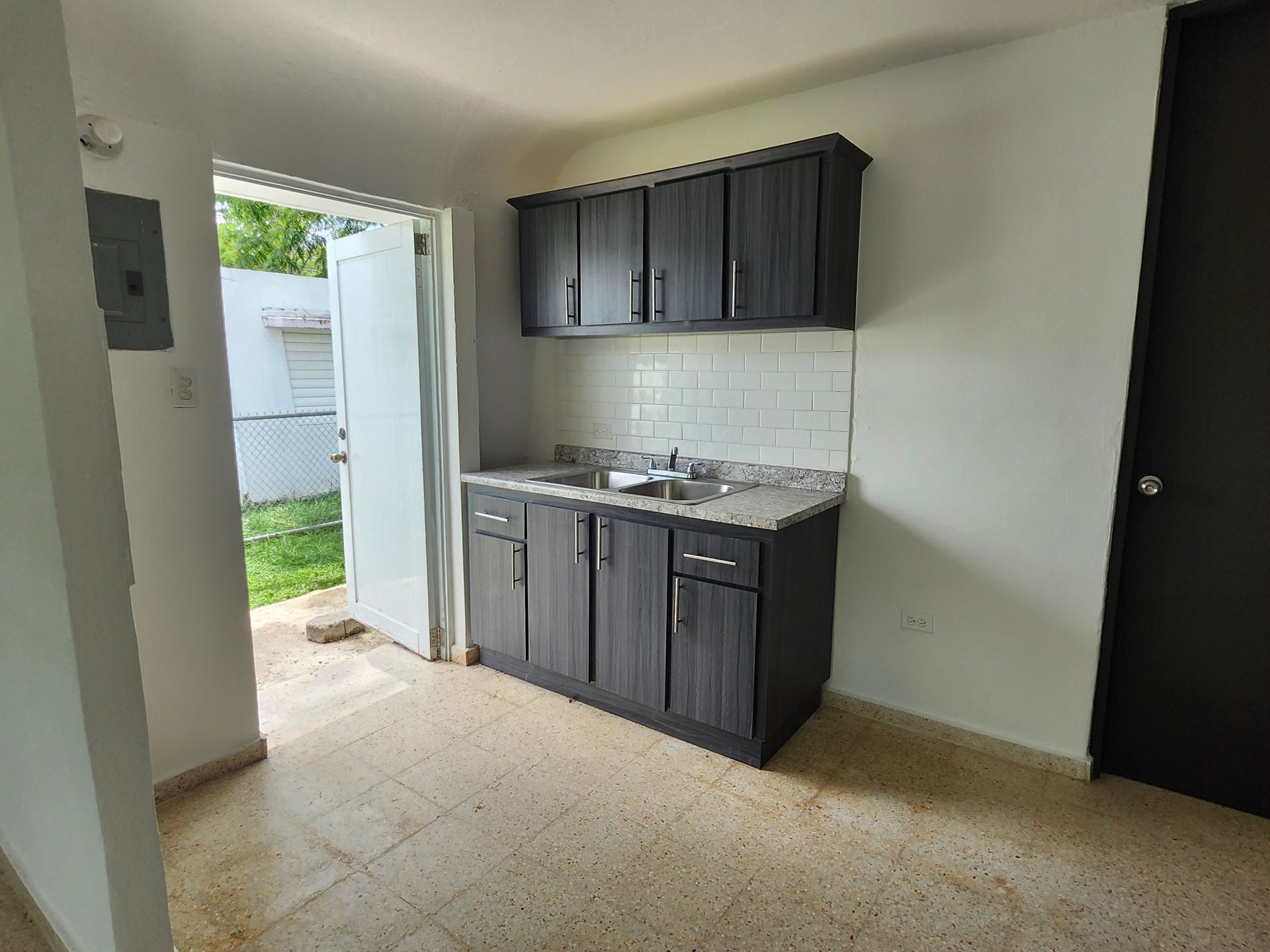 Small kitchenette with dark cabinets and sink in a bare room, open doorway to outside.
