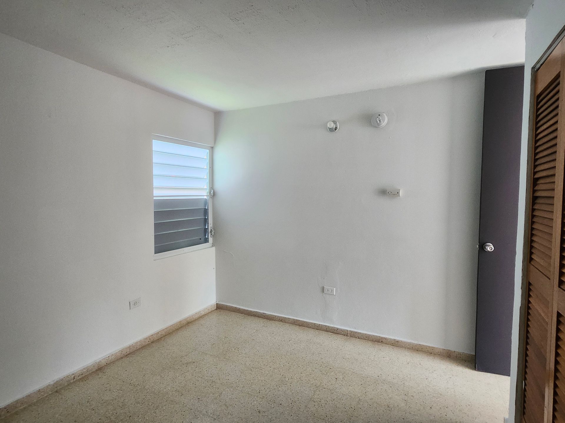 Empty beige bedroom with white walls, small window, and louvered closet doors.