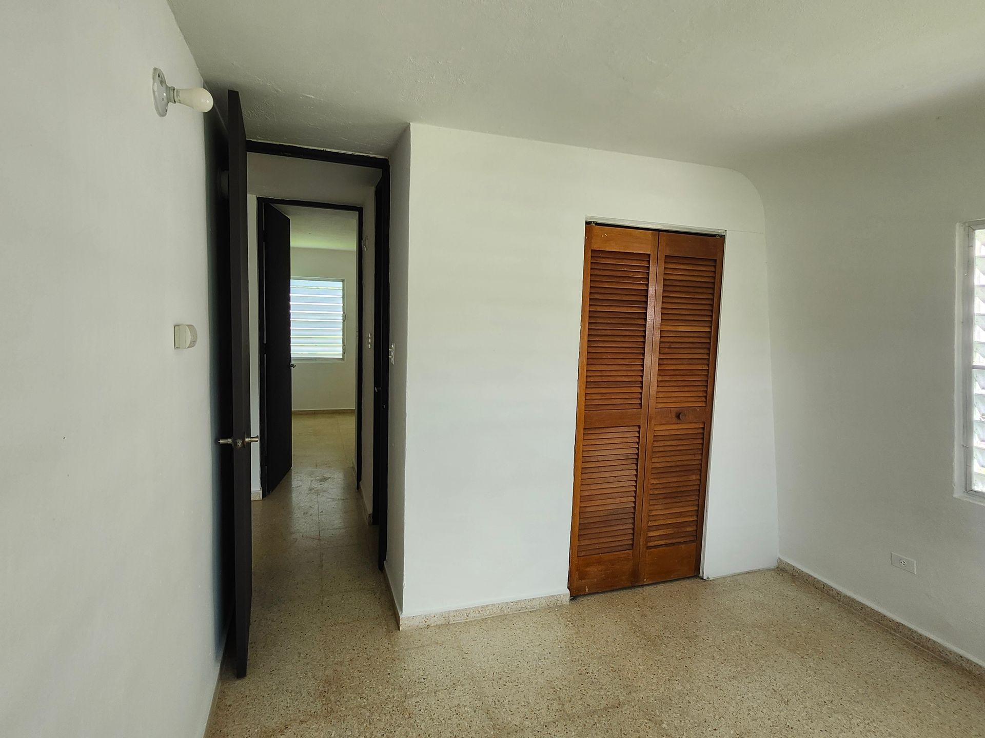Empty room with white walls, speckled floor, open doorway, and wooden louvered closet doors.