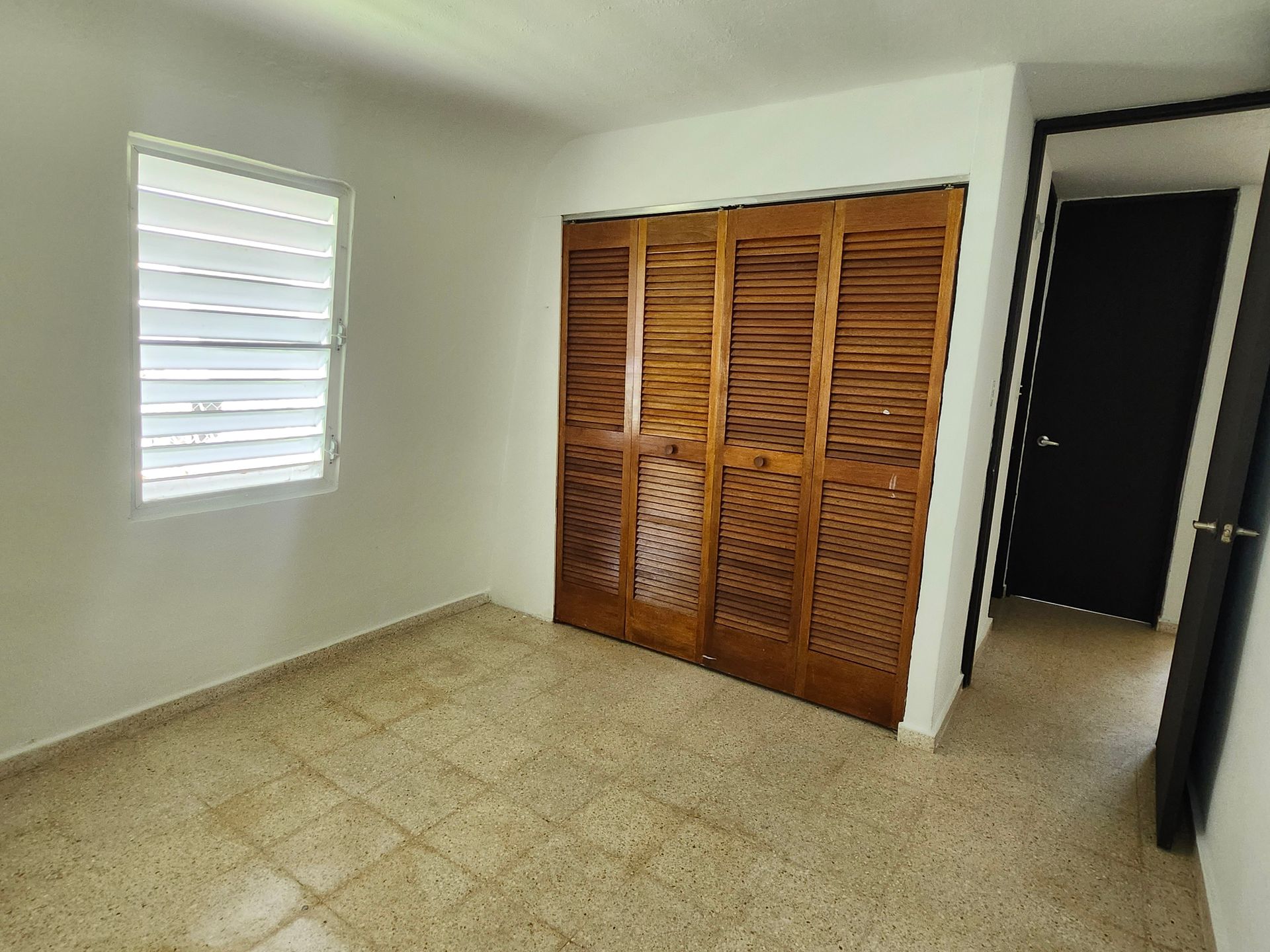 Empty bedroom with beige tile floor, white walls, louvered closet doors, and an open doorway.