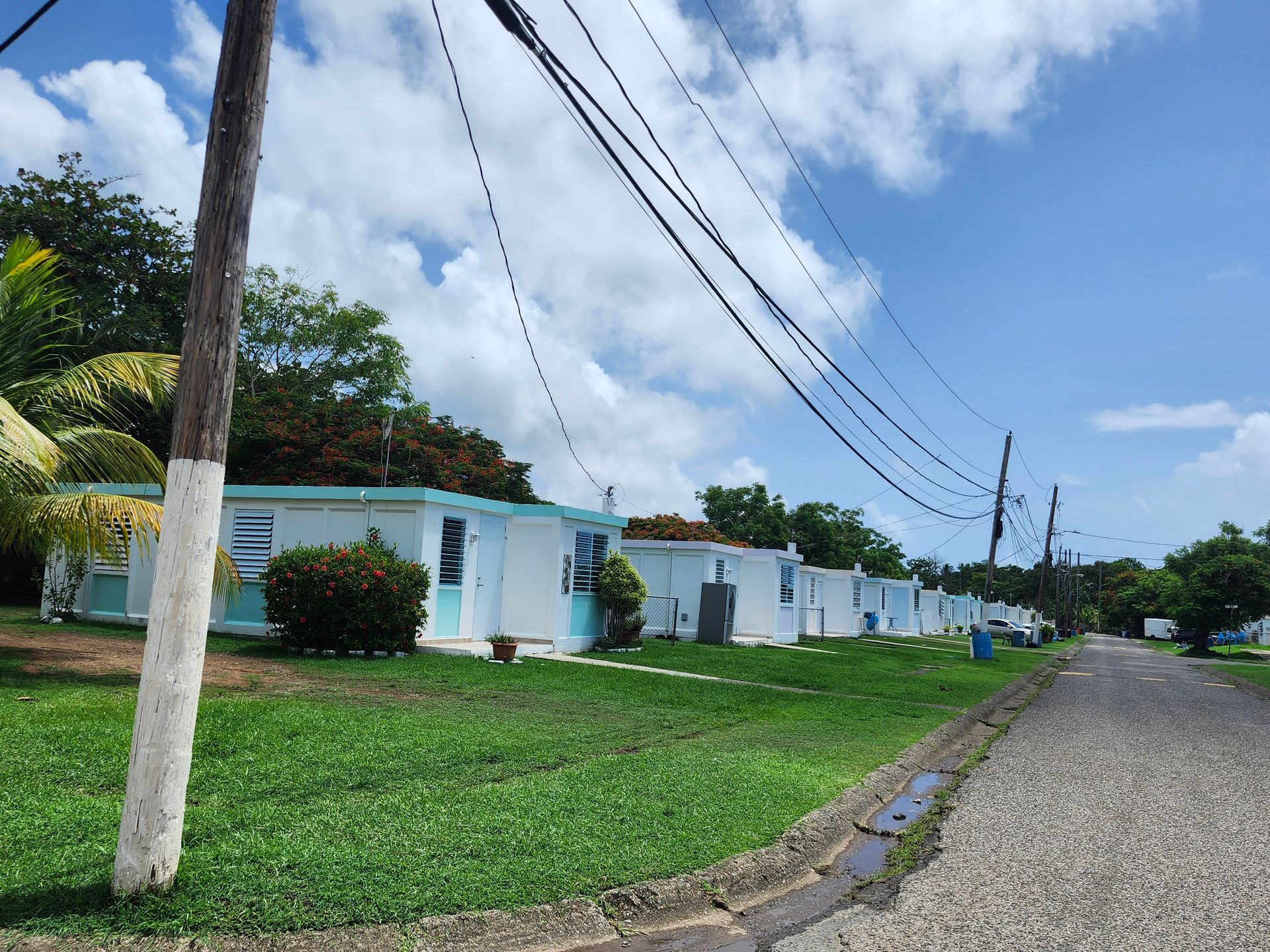 Quiet street with white houses, green lawn, and power lines under a partly cloudy blue sky